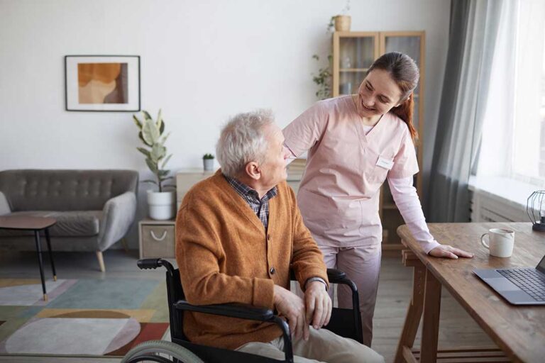 An older adult and family member sit together in a bright assisted living room with photos and personal belongings.